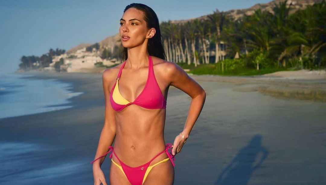 Woman in pink and yellow bikini stands on a beach with palm trees and buildings in the background on a sunny day.