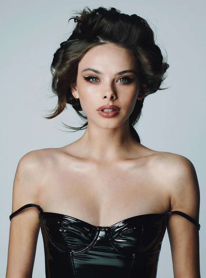Portrait of a beautiful woman with dark hair styled in curlers, wearing a black shiny strapless top against a neutral background.