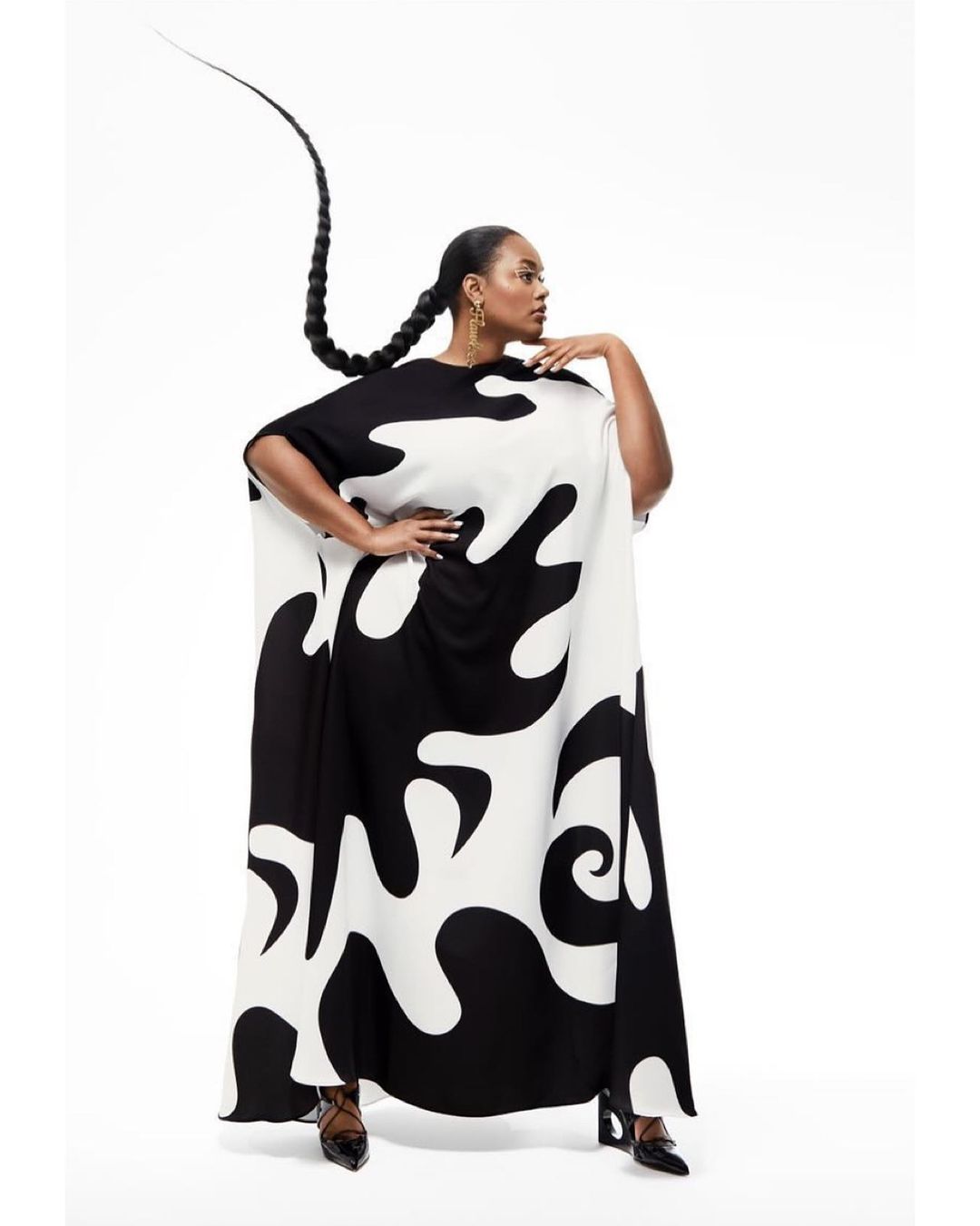 A Black woman in a striking black and white dress poses in a studio, her long braided hair adding a dramatic flair against a bright white backdrop.