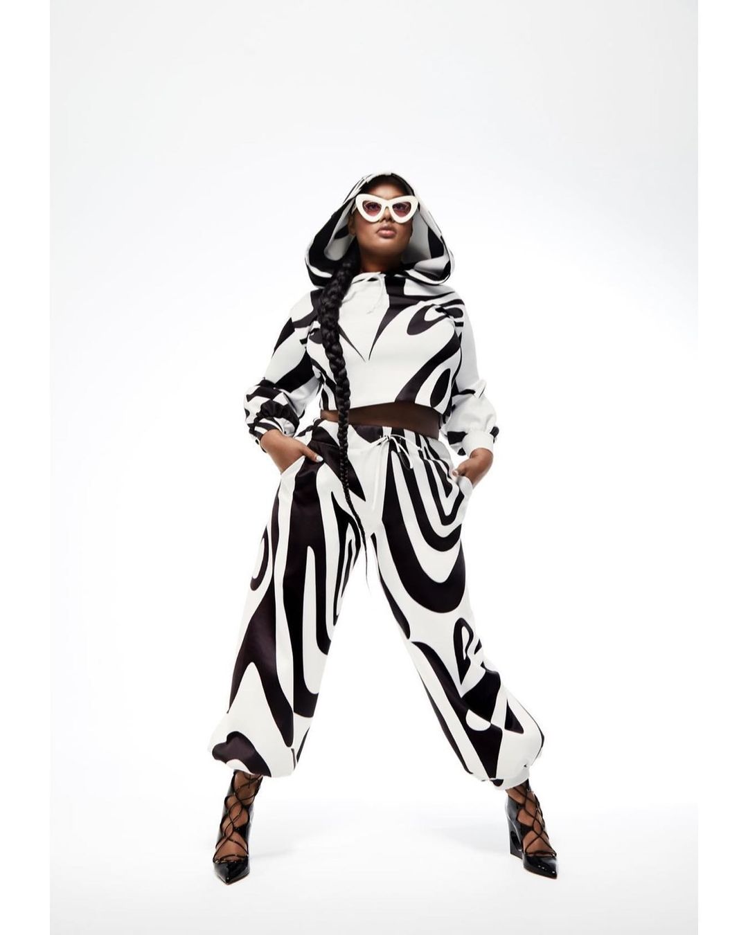 Woman wearing a black and white patterned hooded sweatshirt and pants, along with white sunglasses and black heels poses in a white photo studio.