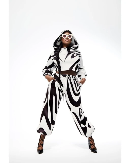 Woman wearing a black and white patterned hooded sweatshirt and pants, along with white sunglasses and black heels poses in a white photo studio.