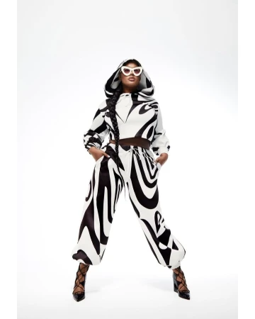 Woman wearing a black and white patterned hooded sweatshirt and pants, along with white sunglasses and black heels poses in a white photo studio.