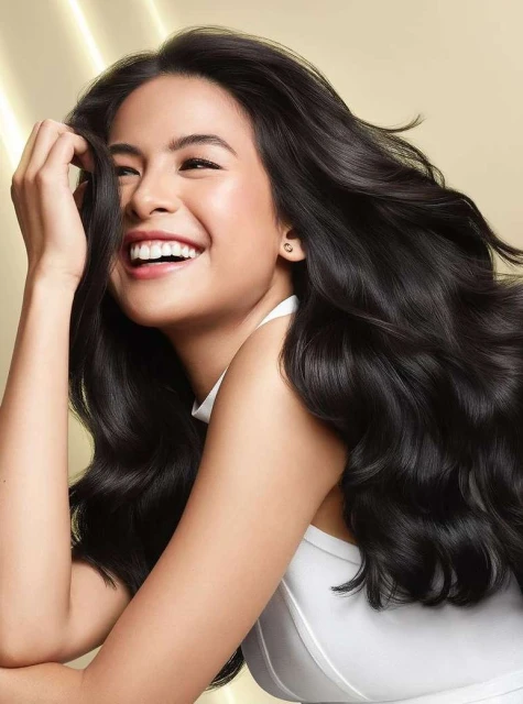 Smiling Asian woman with long, dark, wavy hair against a soft, light-gold background, wearing a white top, studio lighting.