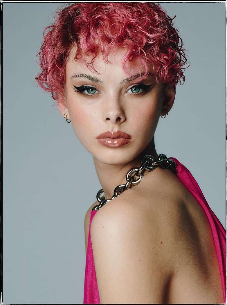 Close-up portrait of a young adult female with pink curly hair, glossy lips, and bold eyeliner, wearing a chain necklace and pink top.