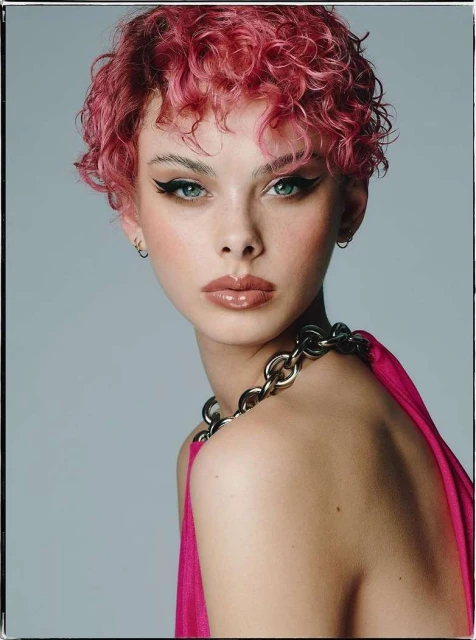 Close-up portrait of a young adult female with pink curly hair, glossy lips, and bold eyeliner, wearing a chain necklace and pink top.