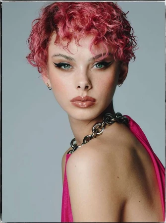 Close-up portrait of a young adult female with pink curly hair, glossy lips, and bold eyeliner, wearing a chain necklace and pink top.