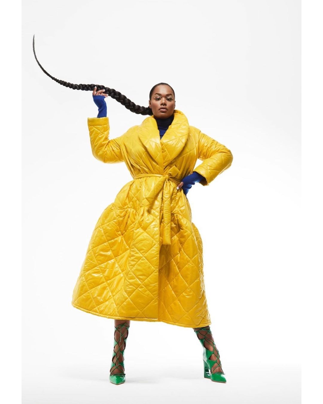 Fashionable woman in a yellow quilted coat, blue top and green high heels, posing in a photo studio with a long black braid.