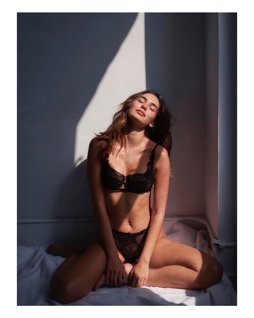 Young woman in black lace lingerie sits with legs crossed against a white wall with natural sunlight creating shadows in a studio setting.