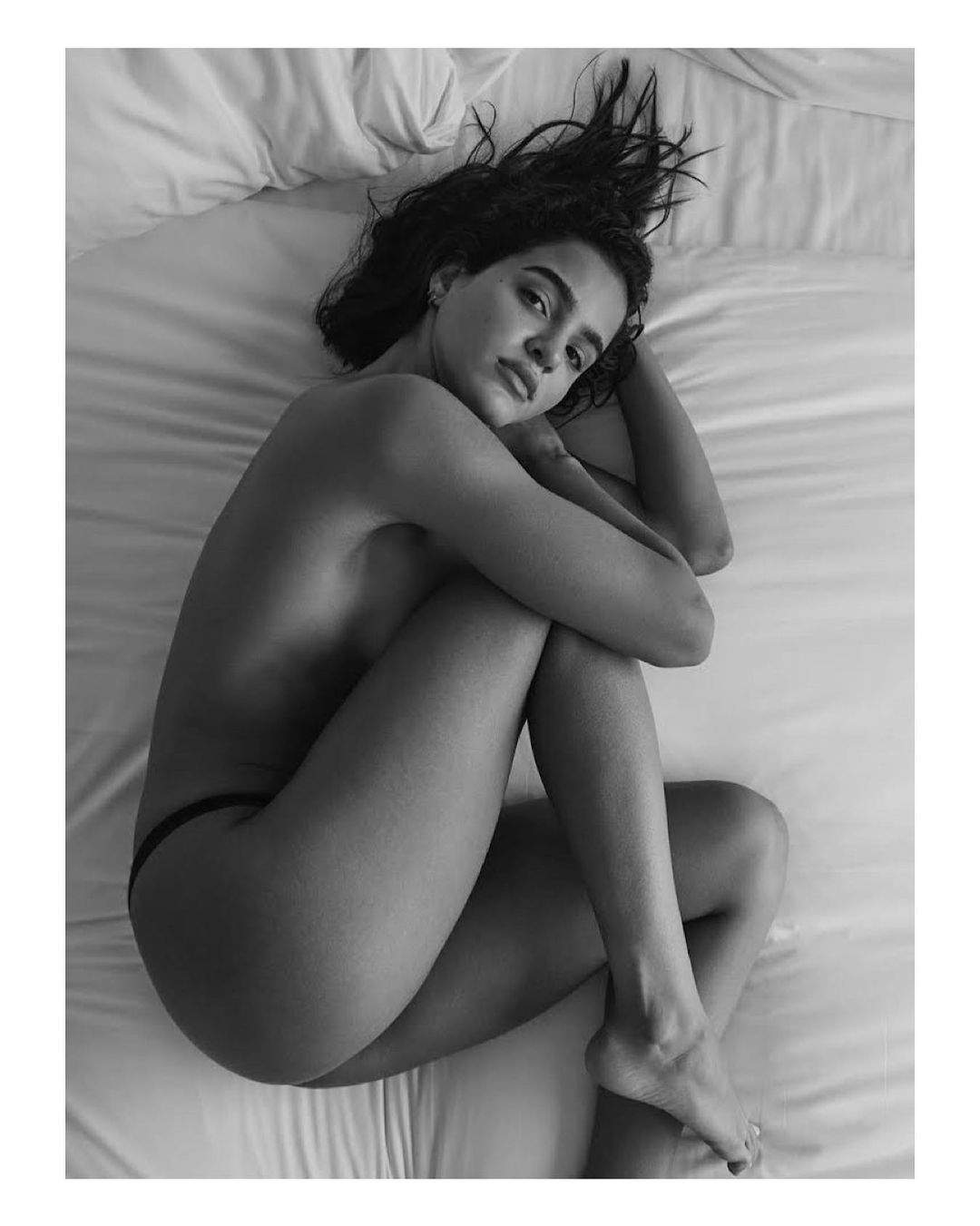 Black and white portrait of an adult woman lying on a bed. She is hugging her knees and looking at the camera with neutral expression.