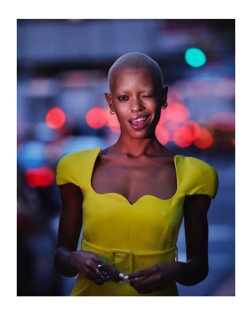 Woman with short blonde hair winking in a yellow dress, posing against an out-of-focus urban background with bokeh, evening lighting.