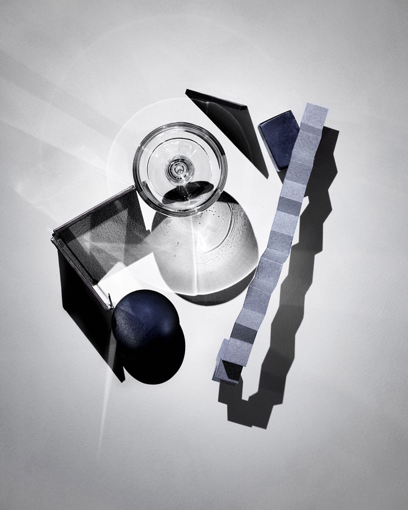 Overhead shot of arranged transparent glass, opaque blue sphere, and geometric paper strips on a white background with shadows.