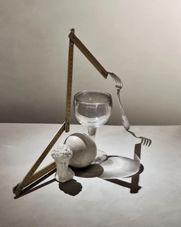Artistic still life: Bust of sculpture, wooden ball, drinking glass, fork, and folding ruler on a gray surface with dramatic shadows.