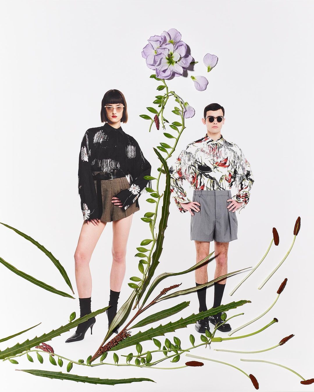 Two people in fashion shot with decorative greenery and floral accents. Man and woman wearing stylish outfits on a white background.