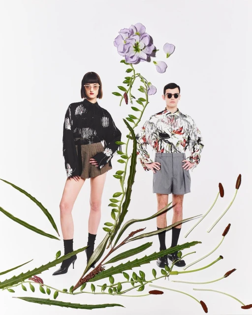 Two people in fashion shot with decorative greenery and floral accents. Man and woman wearing stylish outfits on a white background.