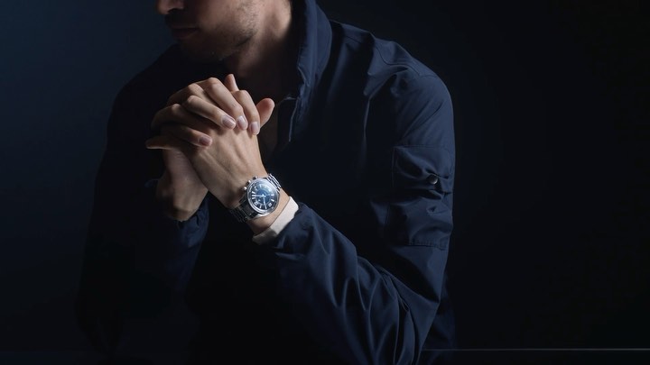 Man with crossed hands wearing a wristwatch in a dimly lit indoor setting. Dark navy jacket, and light skin tone.