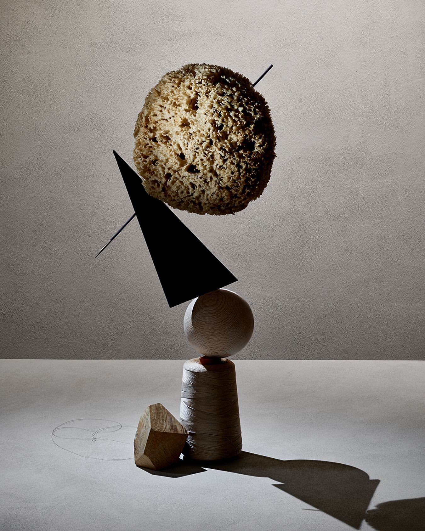 Abstract still life of stacked geometric shapes, including a sea sponge, black triangle, and wooden sphere, set against a gray background.