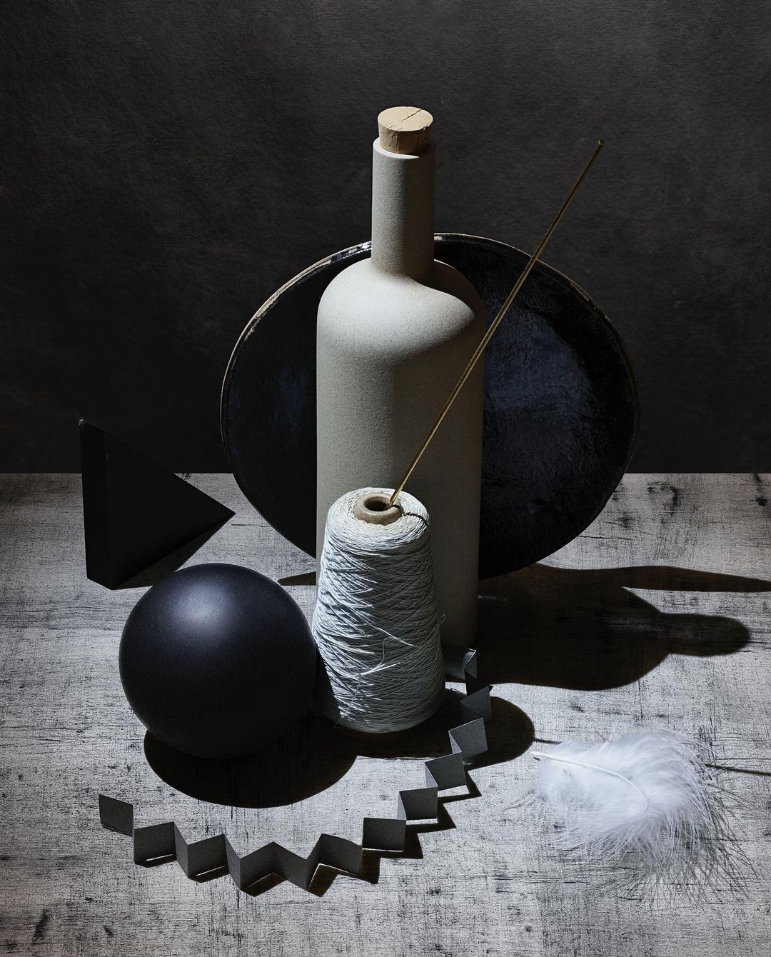 Still life photography showcasing a bottle, spool of thread, ball, feather, and geometric shapes on a wooden surface against a dark background.