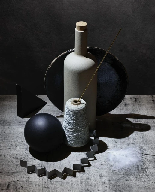Still life photography showcasing a bottle, spool of thread, ball, feather, and geometric shapes on a wooden surface against a dark background.