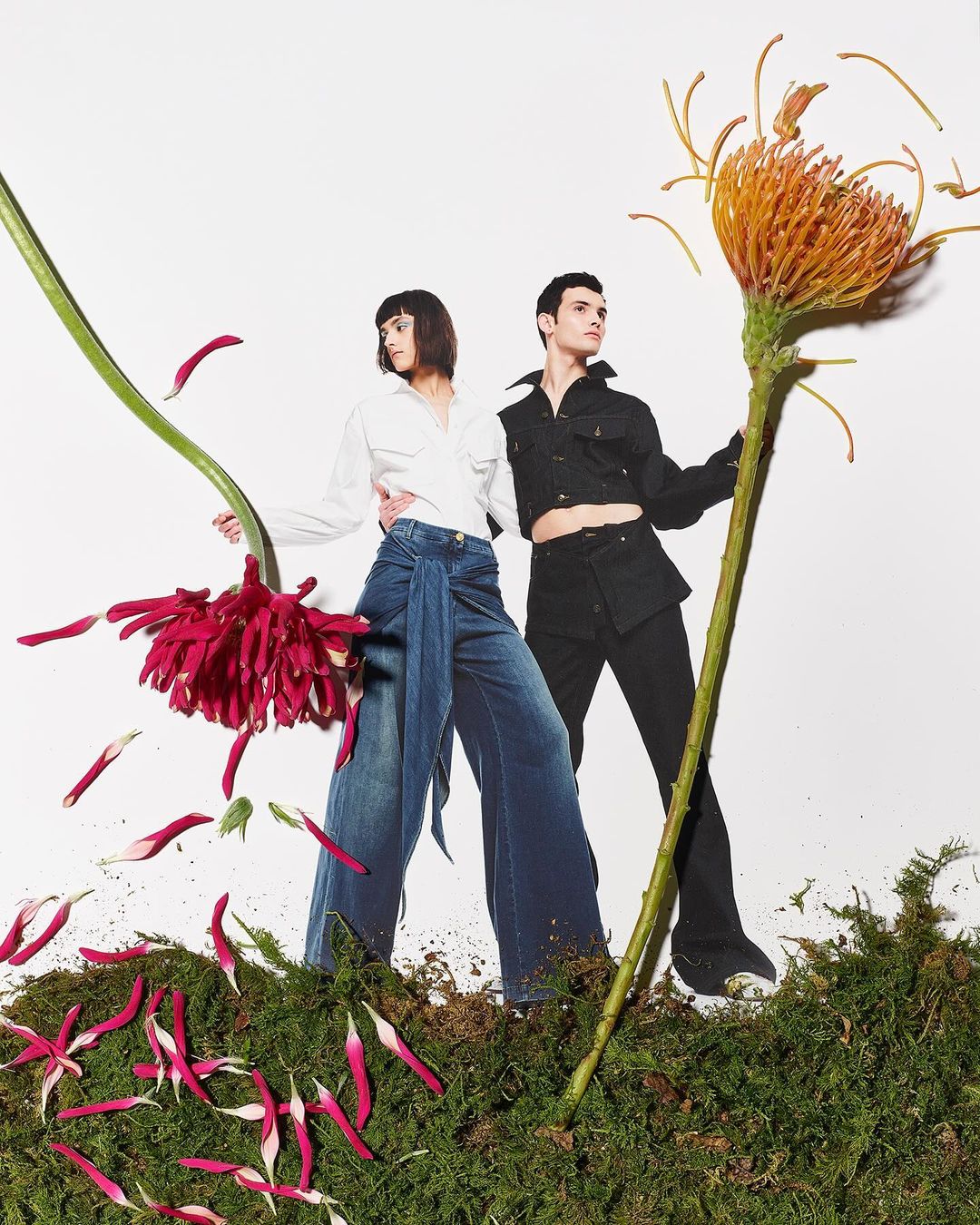 Two models pose among oversized flowers and moss, wearing a white button-down shirt and dark denim jeans and a black jacket with black pants.