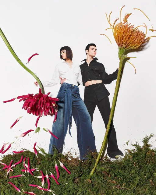 Two models pose among oversized flowers and moss, wearing a white button-down shirt and dark denim jeans and a black jacket with black pants.