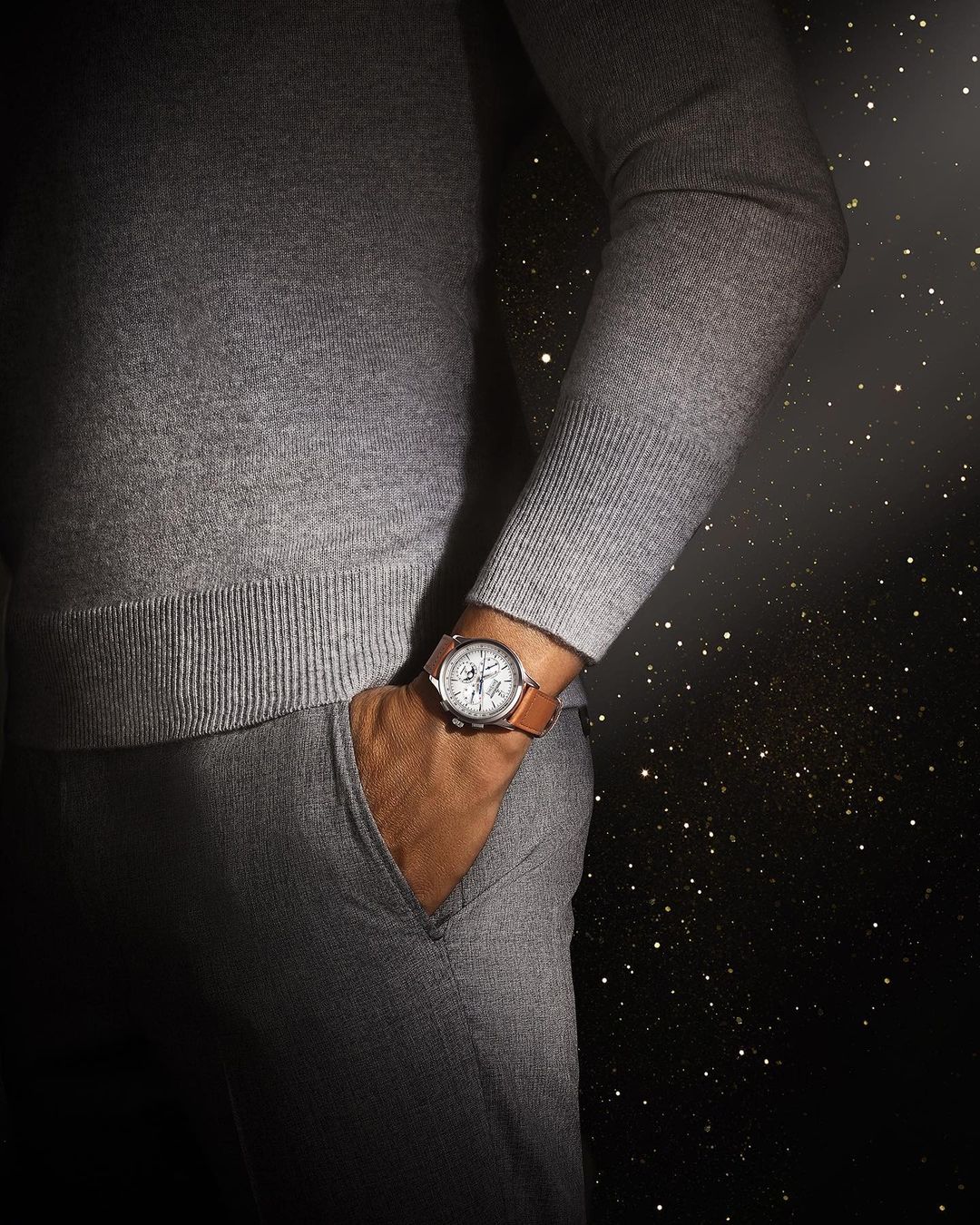 Person wearing a gray sweater and pants, with a wristwatch on their left wrist, standing against a dark backdrop with bokeh.