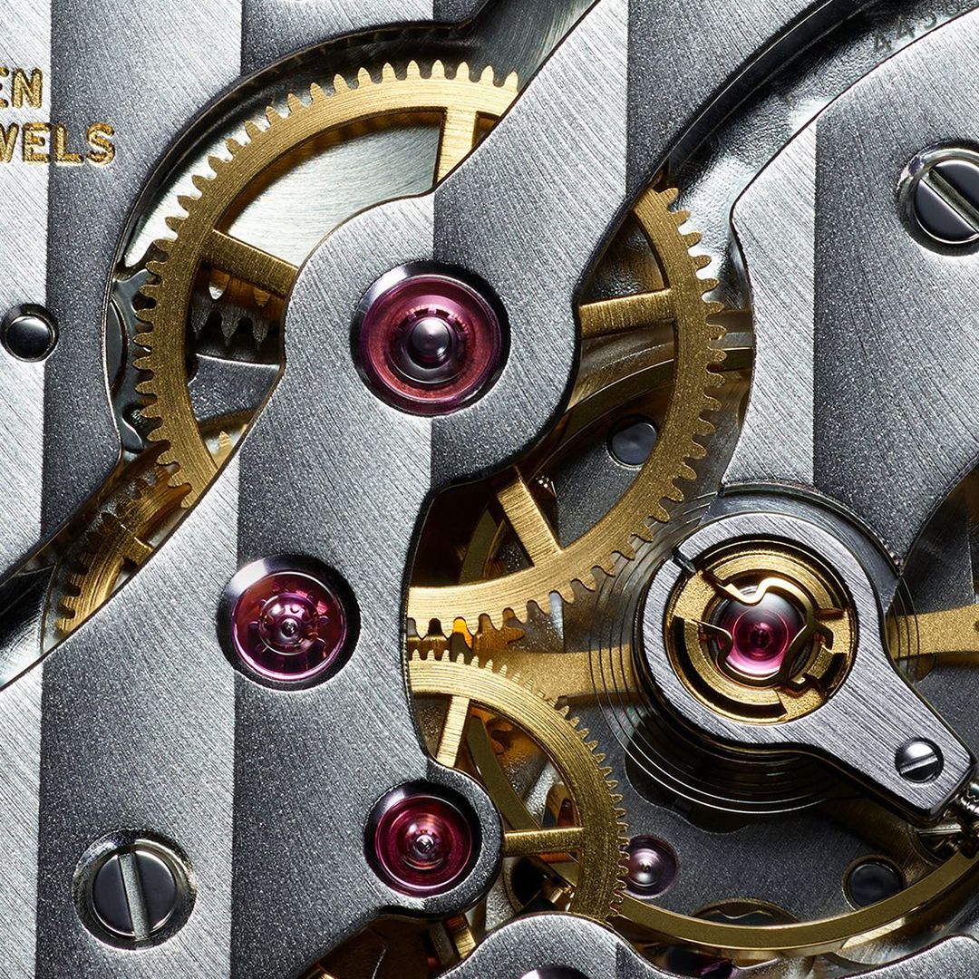 Close-up of intricate watch mechanism featuring golden gears, screws, and jewels against a textured silver metal background.