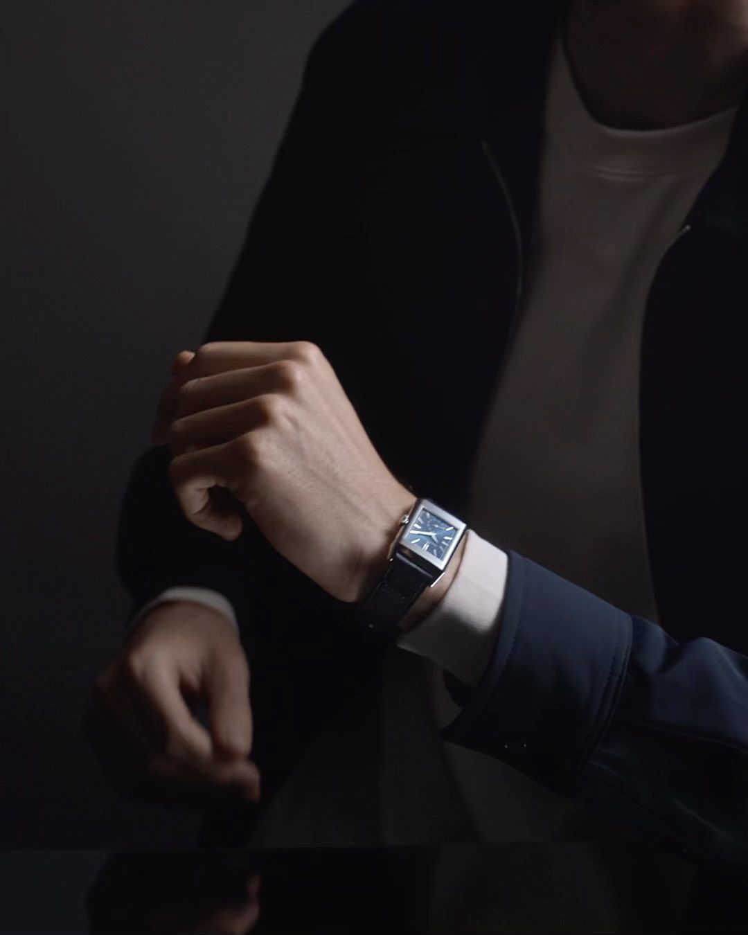 Close-up on an adult's wrist wearing a silver wristwatch, visible next to a black jacket sleeve and a black glossy surface.