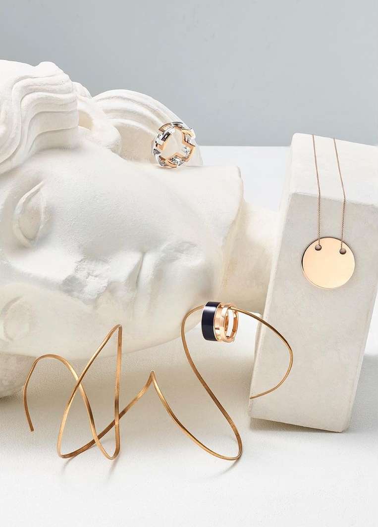 Still life of gold jewelry, featuring rings and a necklace, arranged on white blocks and a classical bust against a soft white background.