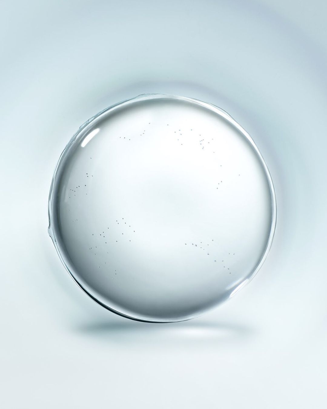 A clear, round droplet or gel sits against a soft, light blue gradient background, highlighted by a central reflection point.