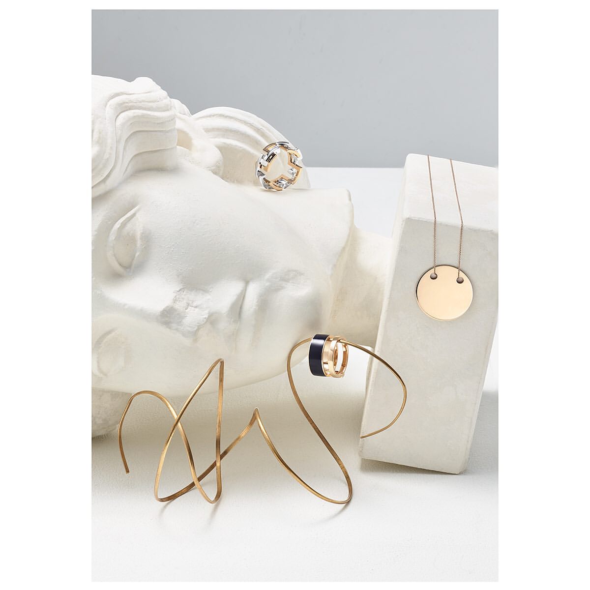 Jewelry product shot featuring gold and silver rings, a gold necklace, and geometric art pieces, set against a white plaster head.