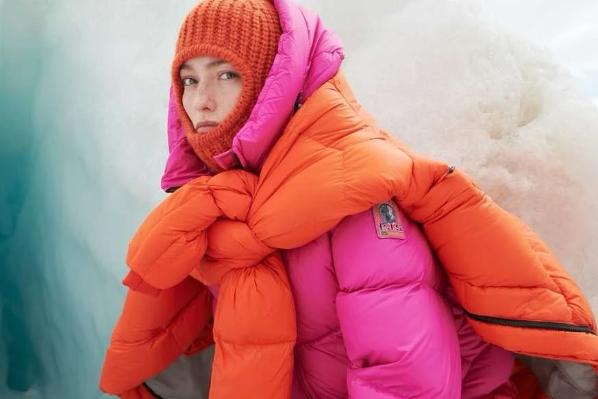 Close-up of a woman wearing a bright orange balaclava and a pink and orange puffer jacket posing in front of a glacier backdrop.