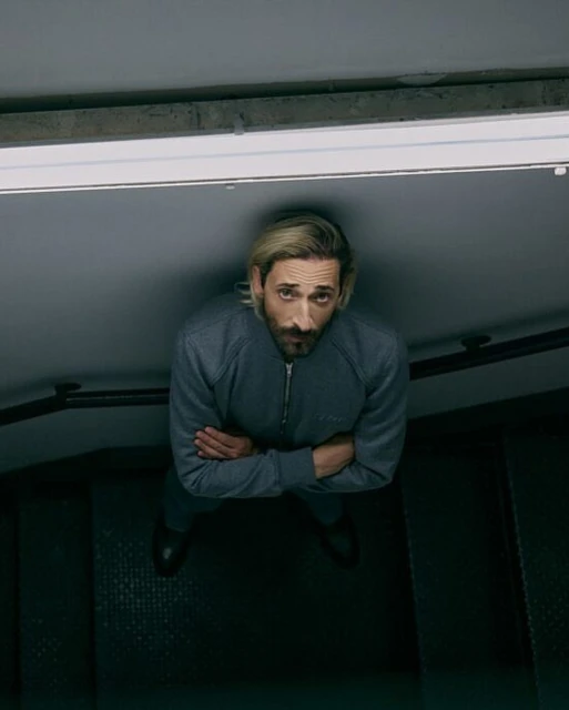 Eye-level view of an adult male with crossed arms wearing a blue zip-up sweatshirt and black dress shoes, looking up at the camera.