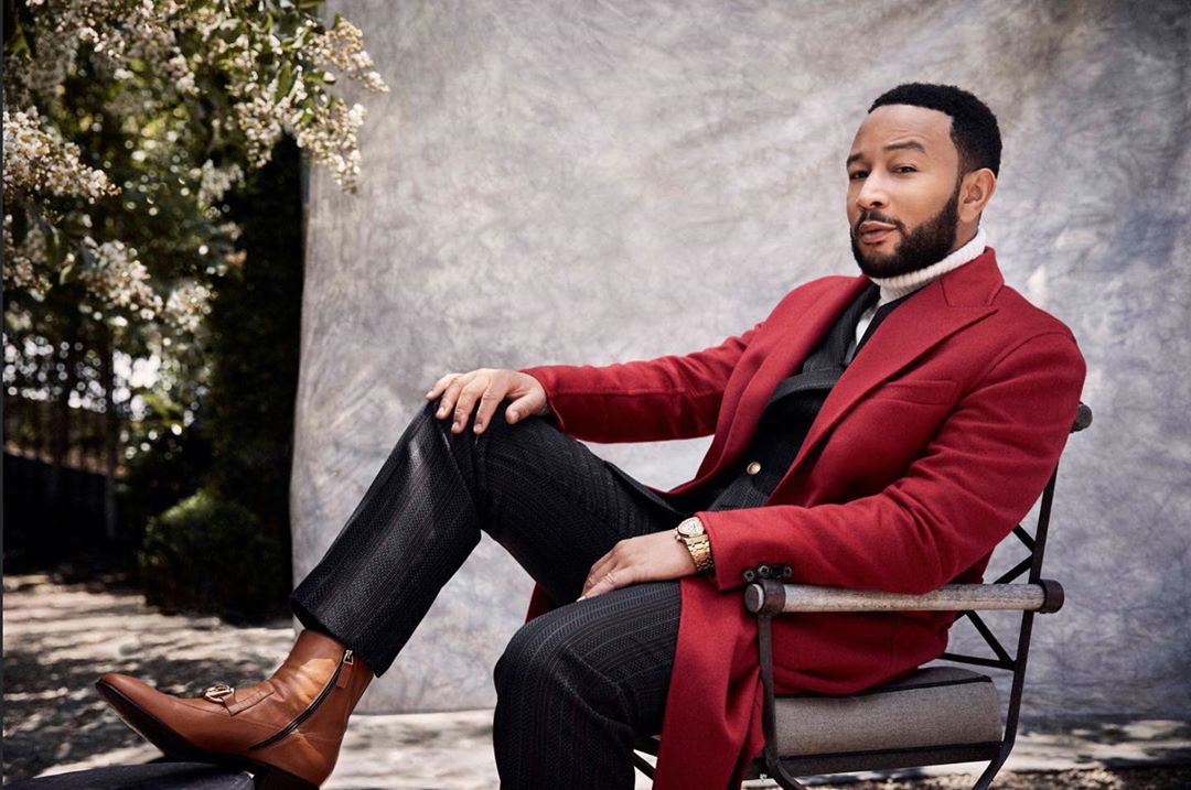Adult male poses in an outdoor portrait, sitting in a chair wearing a red coat, patterned pants, and tan boots against a neutral backdrop.