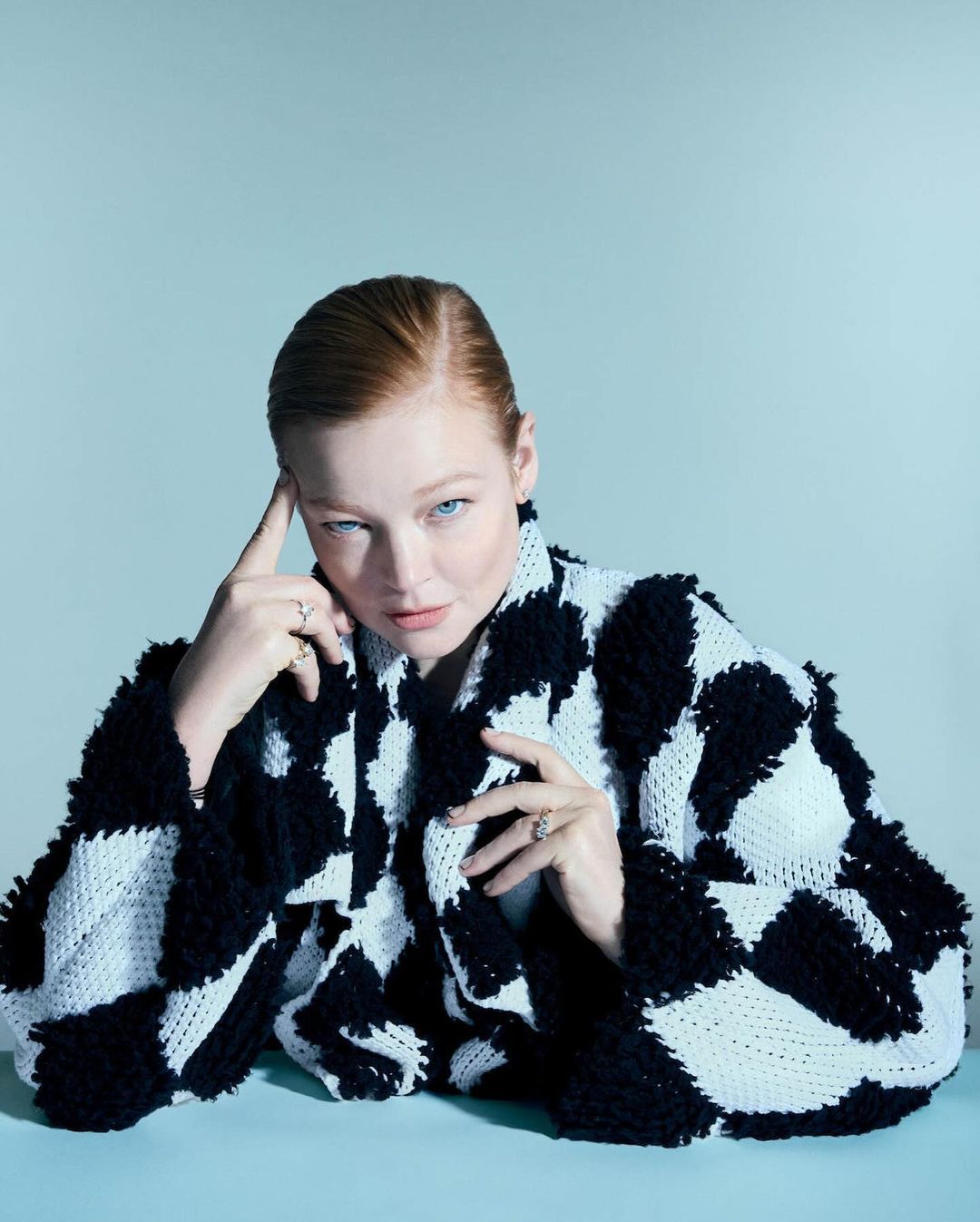 Red-haired woman in a black-and-white checkerboard sweater poses indoors against a light blue background, gazing intently at the camera.