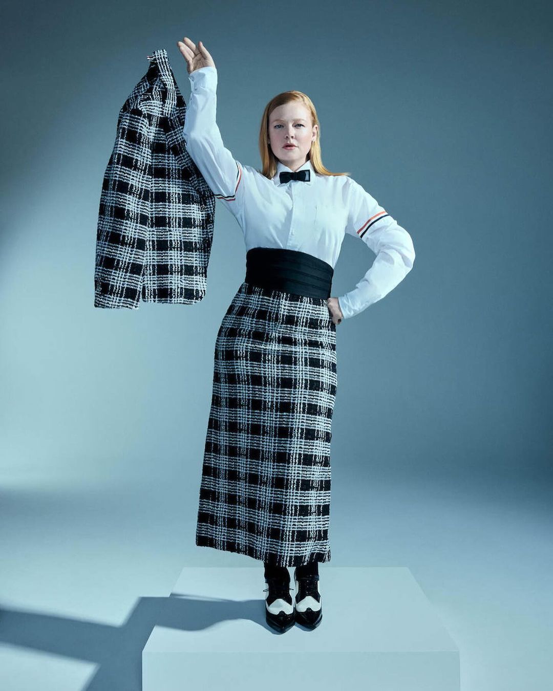 Fashionable woman in black-and-white plaid skirt, white shirt, and black bow tie poses in a photo studio holding a matching jacket.