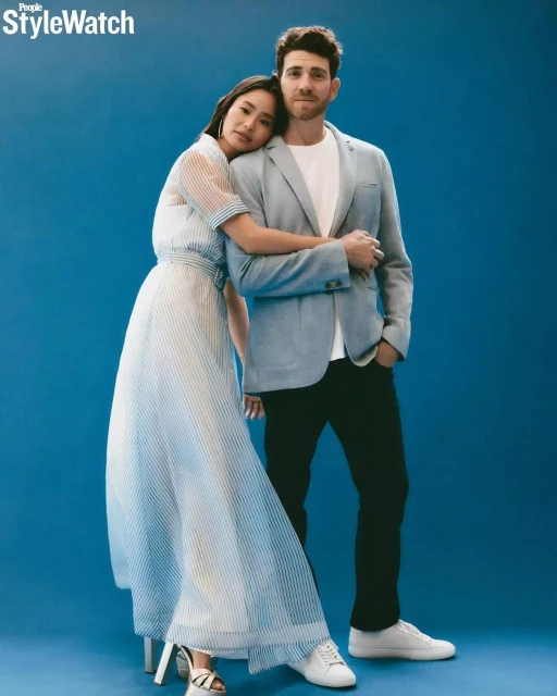 Fashionable couple in studio portrait: woman in striped maxi dress leans on man in blazer against blue background. Modern style and posing.