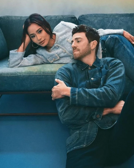 A stylish couple poses indoors on a blue couch. The woman wears a light blue tweed jacket, and the man wears a denim jacket.