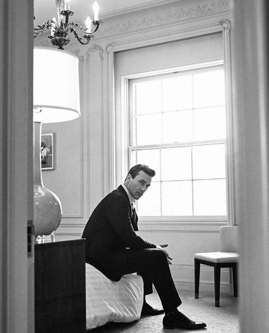 Black and white portrait of an adult man in a dark suit, sitting on a bed in a bright room near a window and a white chair.