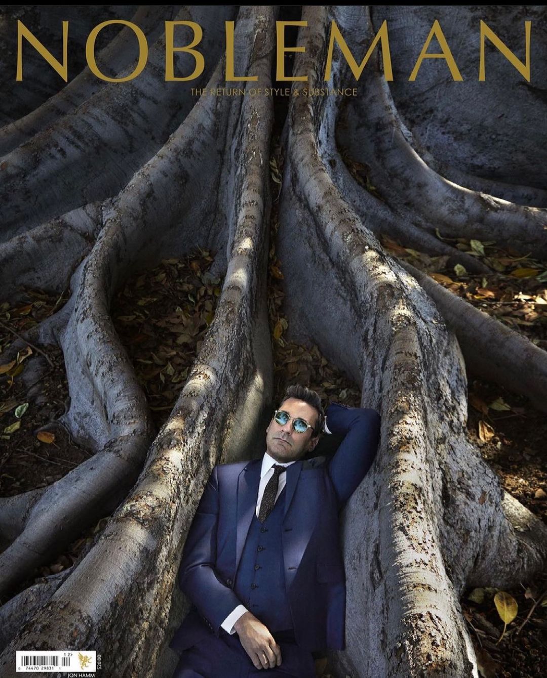 Magazine cover featuring a man in a blue suit reclining among the exposed roots of a large tree outdoors in natural light.