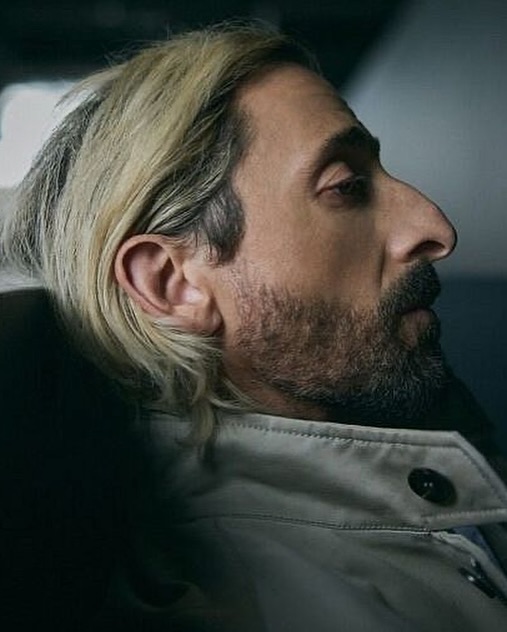 Portrait of a man with dyed blonde hair, a stubbled beard, and a light-colored jacket, captured in a close-up shot with shallow depth of field.