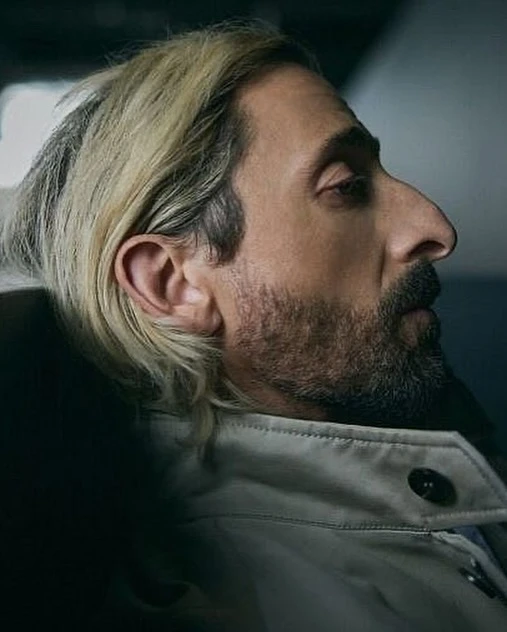 Portrait of a man with dyed blonde hair, a stubbled beard, and a light-colored jacket, captured in a close-up shot with shallow depth of field.