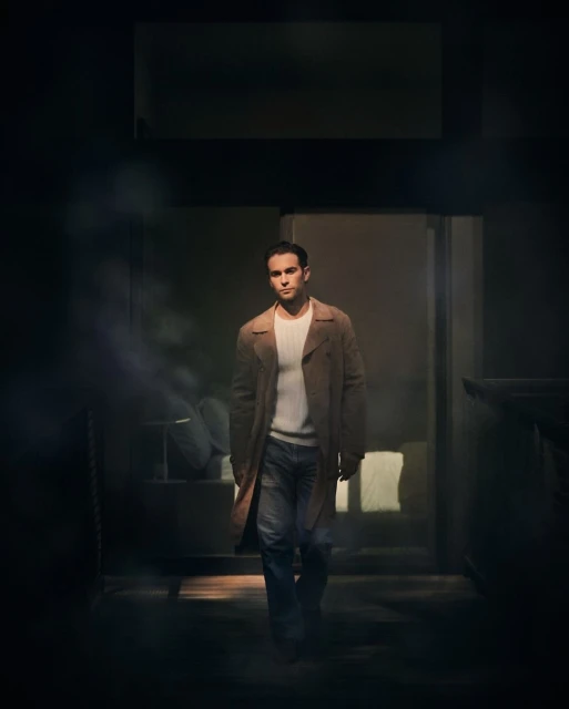 Man in coat walks out of dark doorway with soft light, wearing jeans and white top, medium shot in dimly lit, indoor setting.