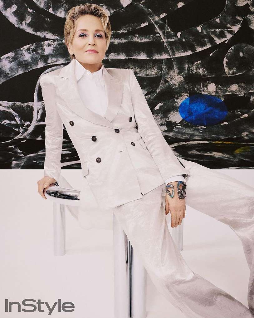 Elegant portrait of Sharon Stone in a white suit and top, seated indoors against an abstract painting with black, white, and blue.
