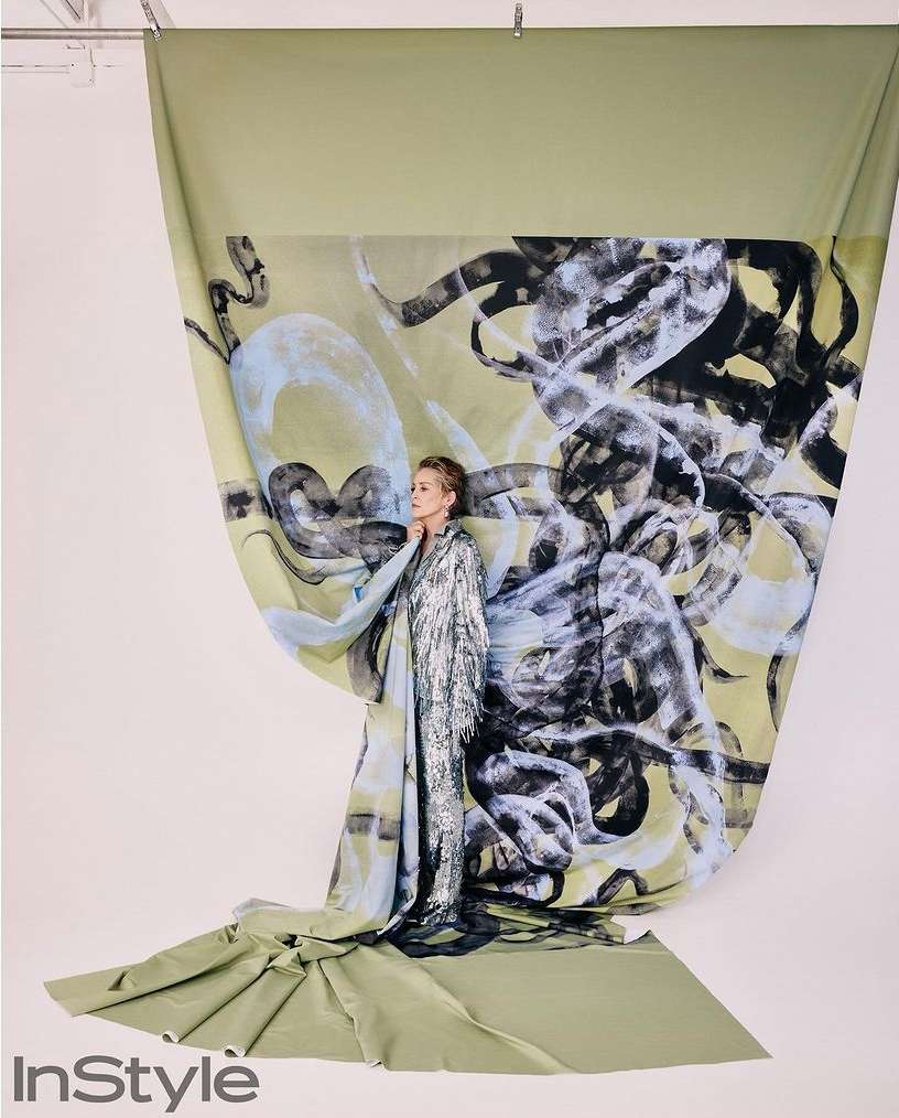 Fashion shot of woman in shiny, fringed dress standing in front of painted backdrop in studio with InStyle logo visible.