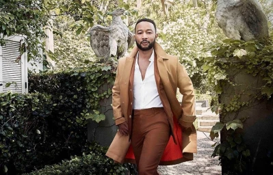 John Legend wearing a camel overcoat, white shirt, and brown trousers outdoors near stone pillars and foliage.