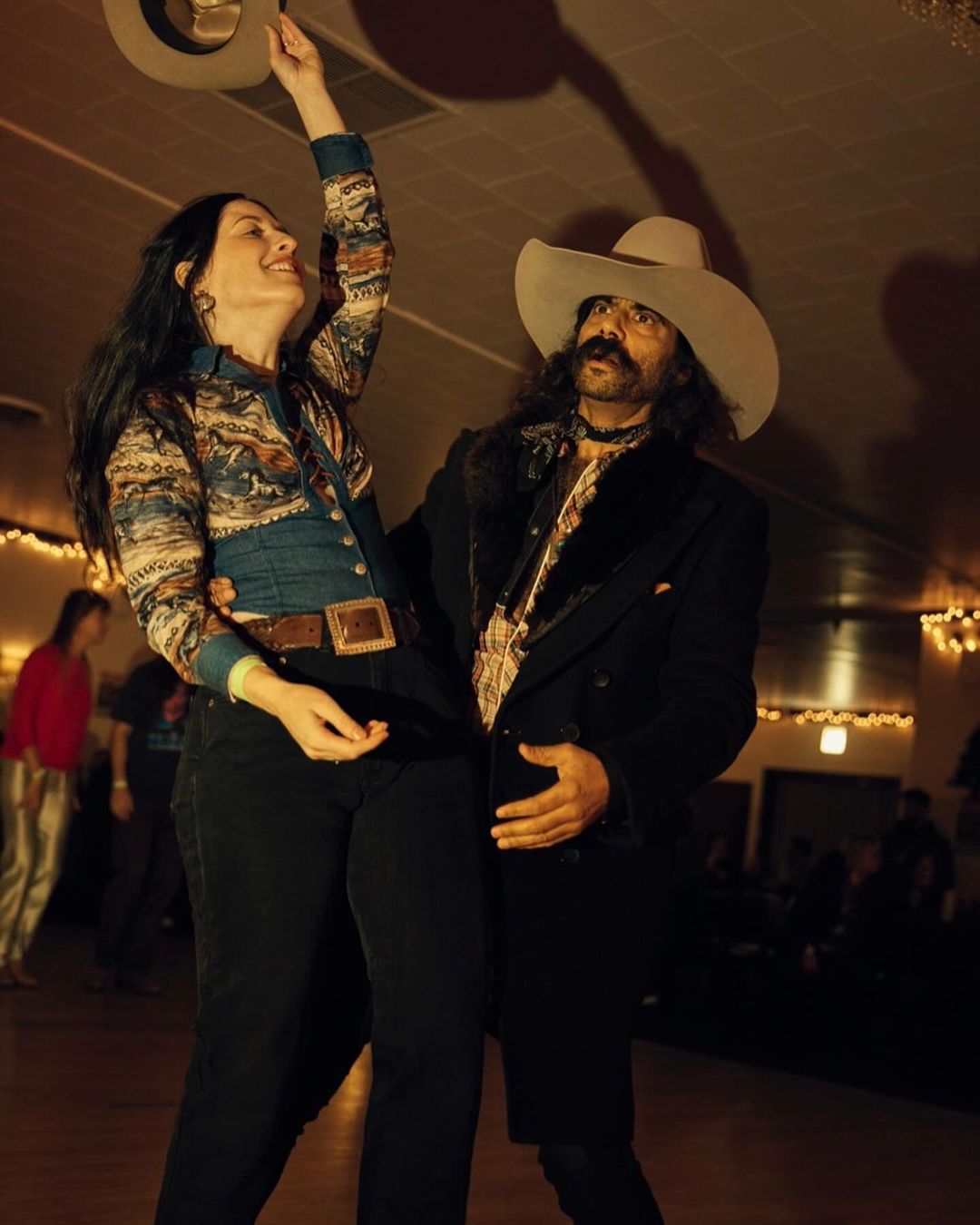A cowboy and woman dancing, the woman is raising a hat. The man in a cowboy hat and black coat smiles at the woman.