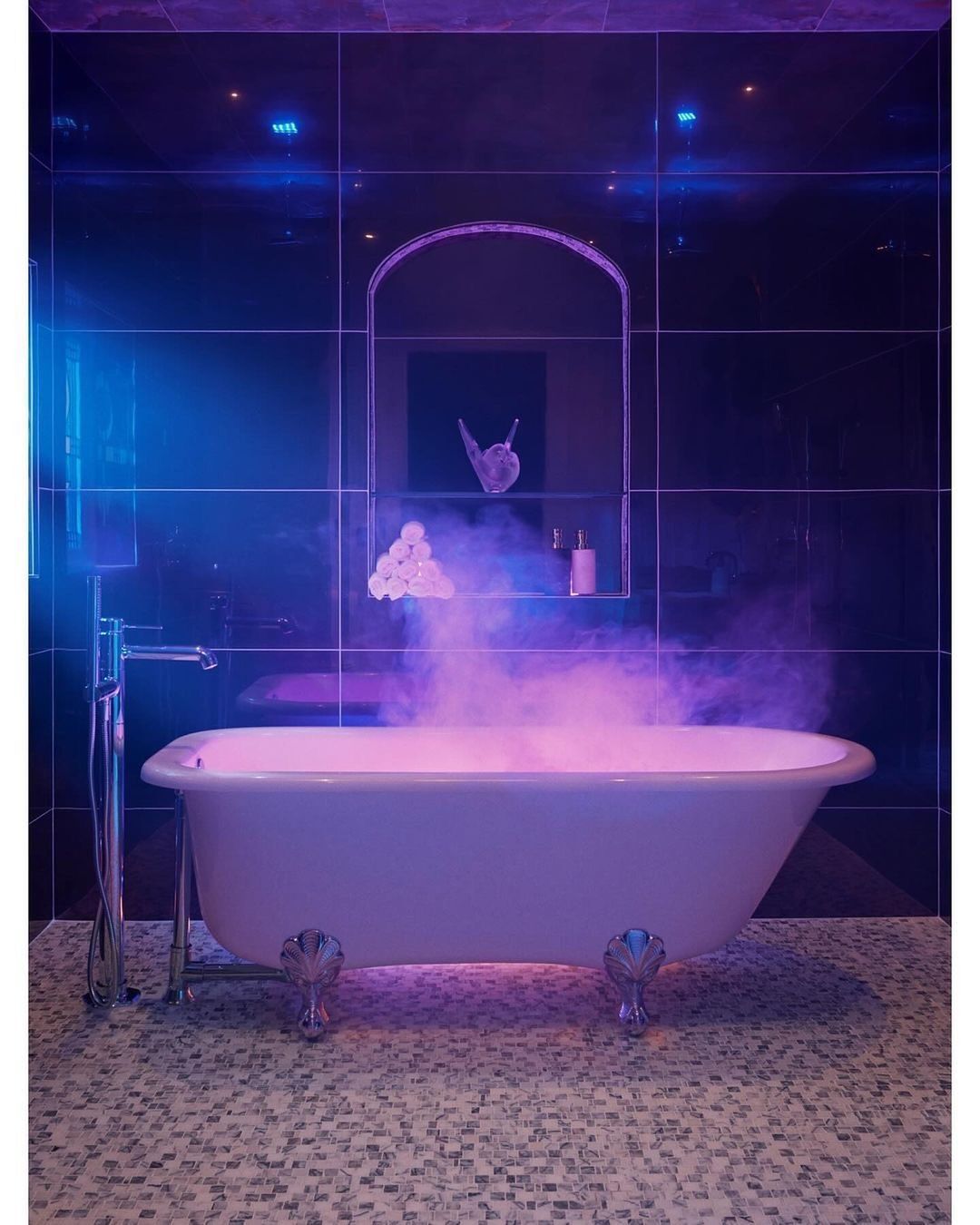 Bathtub in a moody blue and purple bathroom with mosaic tile floor. Bathtub has claw feet and what appears to be steam or fog.