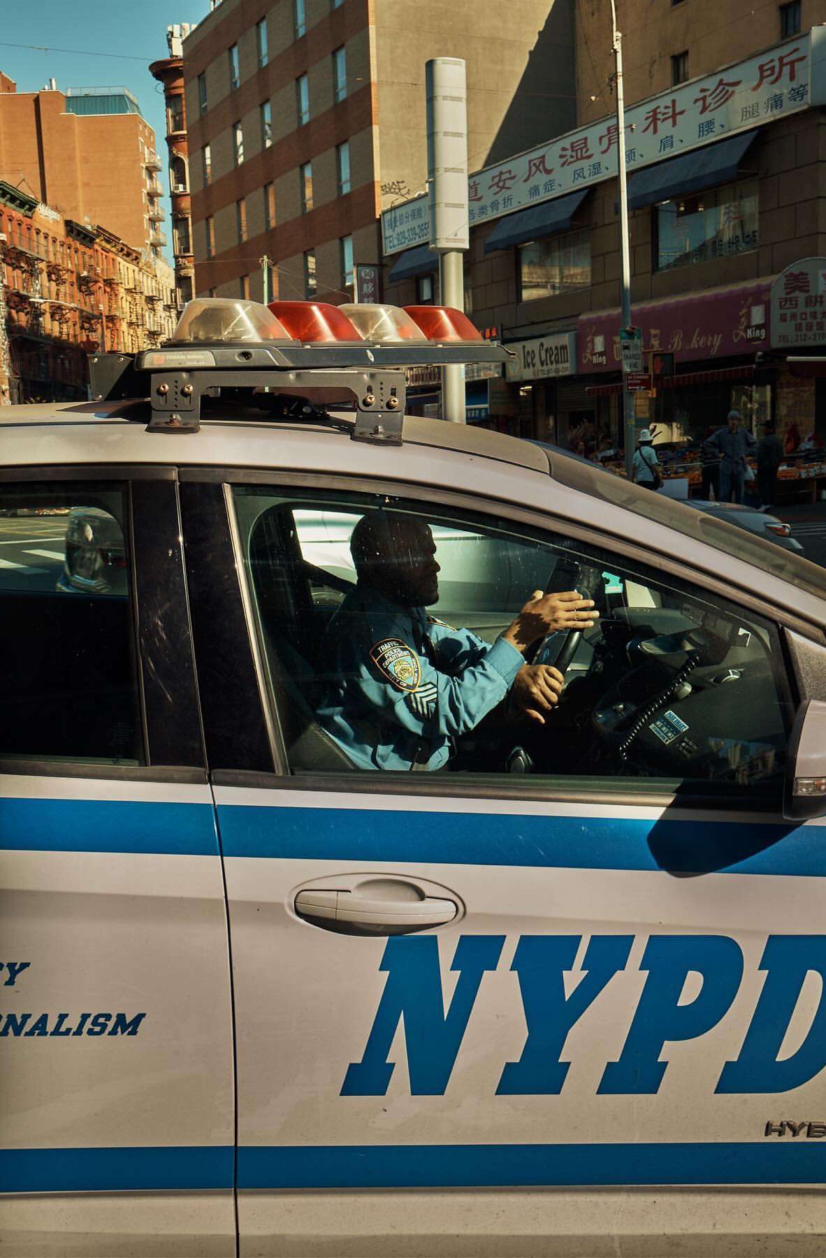 NYPD police car in an urban setting, with a police officer inside, surrounded by city buildings on a sunny day.