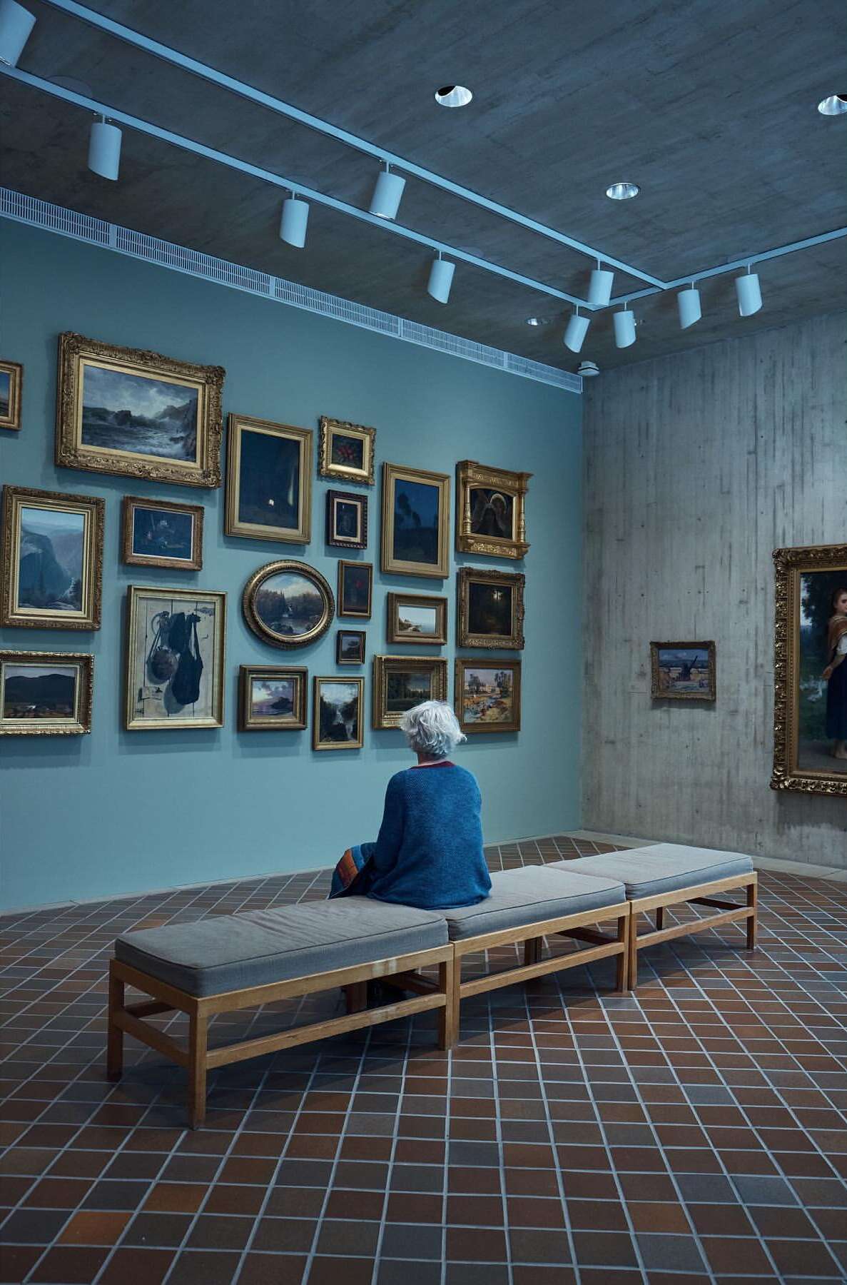 Woman views artwork in a gallery setting. She is sitting on a bench facing a wall filled with paintings in ornate gold frames.