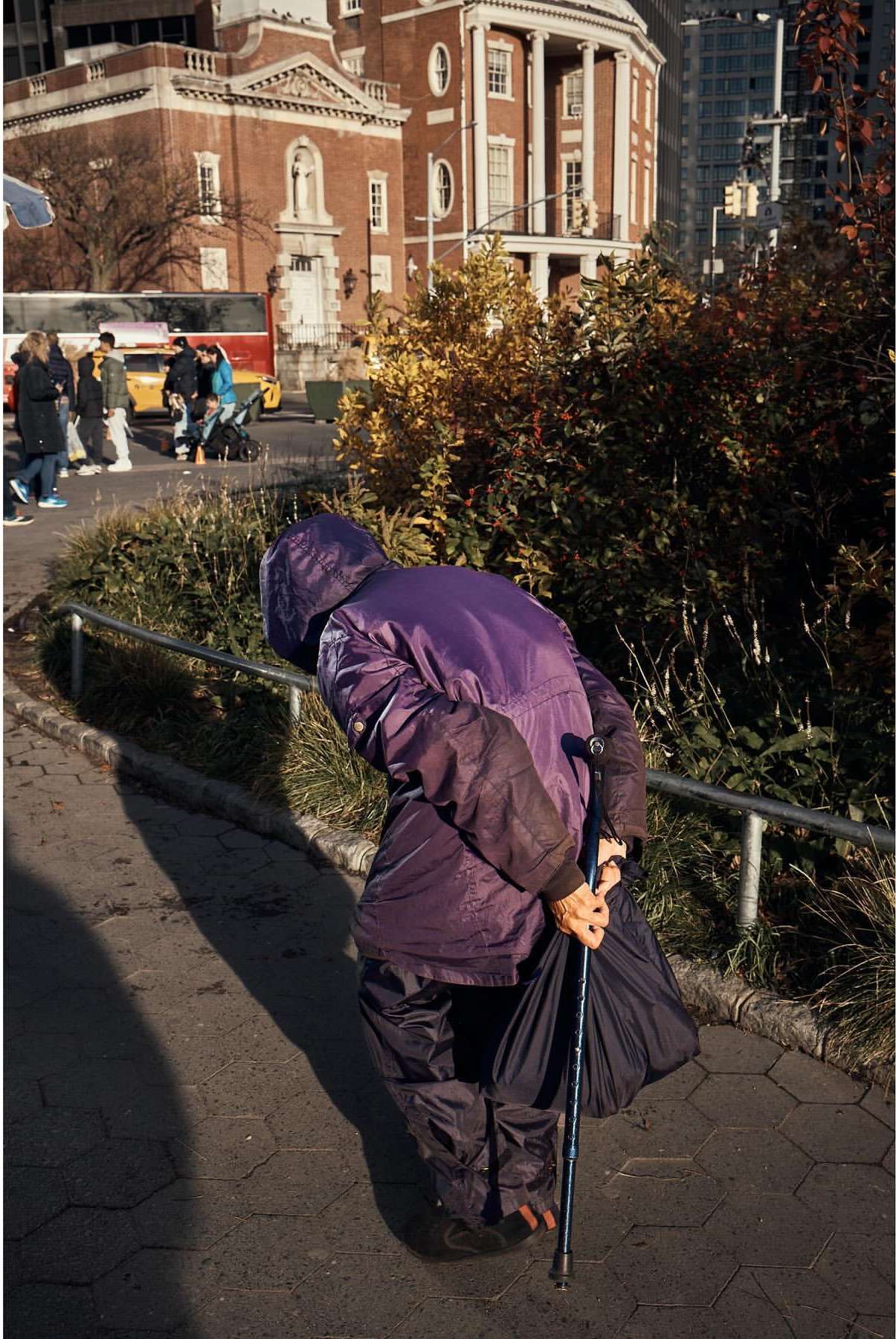 A person with a cane and bag is seen from behind on a stone path with buildings and foliage in the background during daytime.